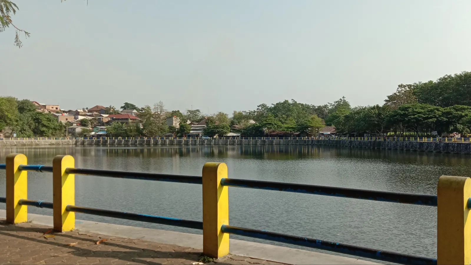 Permukaan air Tambakboyo dan jalur tepian berpagar kuning hitam dengan permukiman serta pepohonan di kejauhan